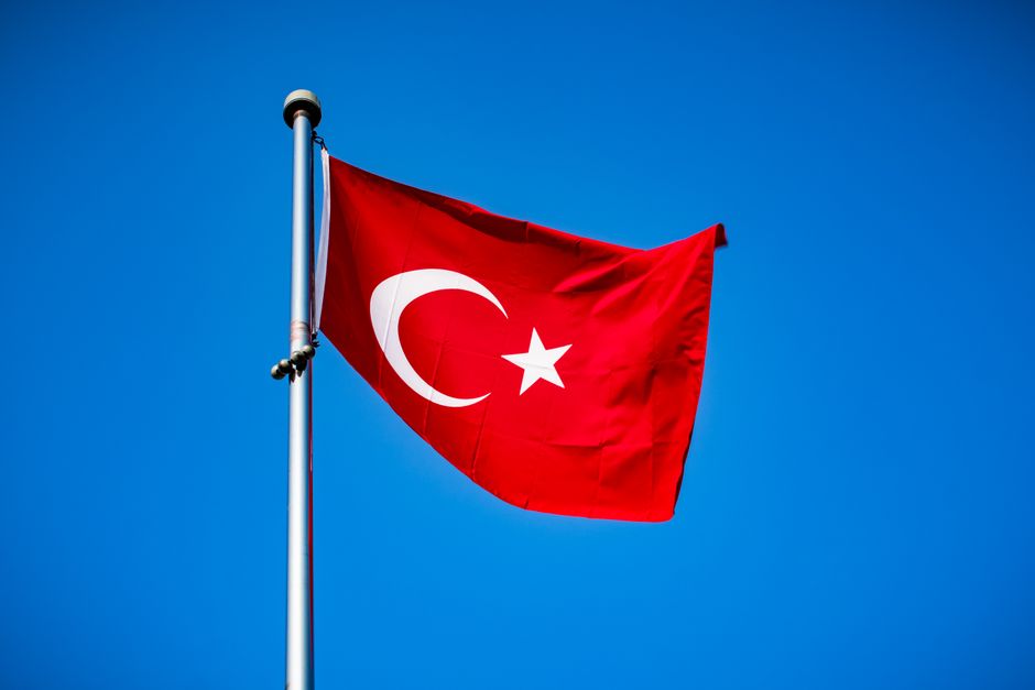 Parlak mavi gökyüzü önünde, hilal ve yıldızla süslü canlı kırmızı Türk bayrağı dalgalanıyor.